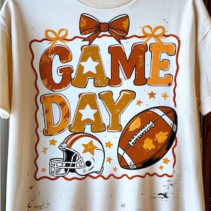 May include: Cream-colored t-shirt featuring a football theme. The design includes the words "GAME DAY" in orange and brown, a football, a helmet, and star accents. A bow and decorative border complete the design.