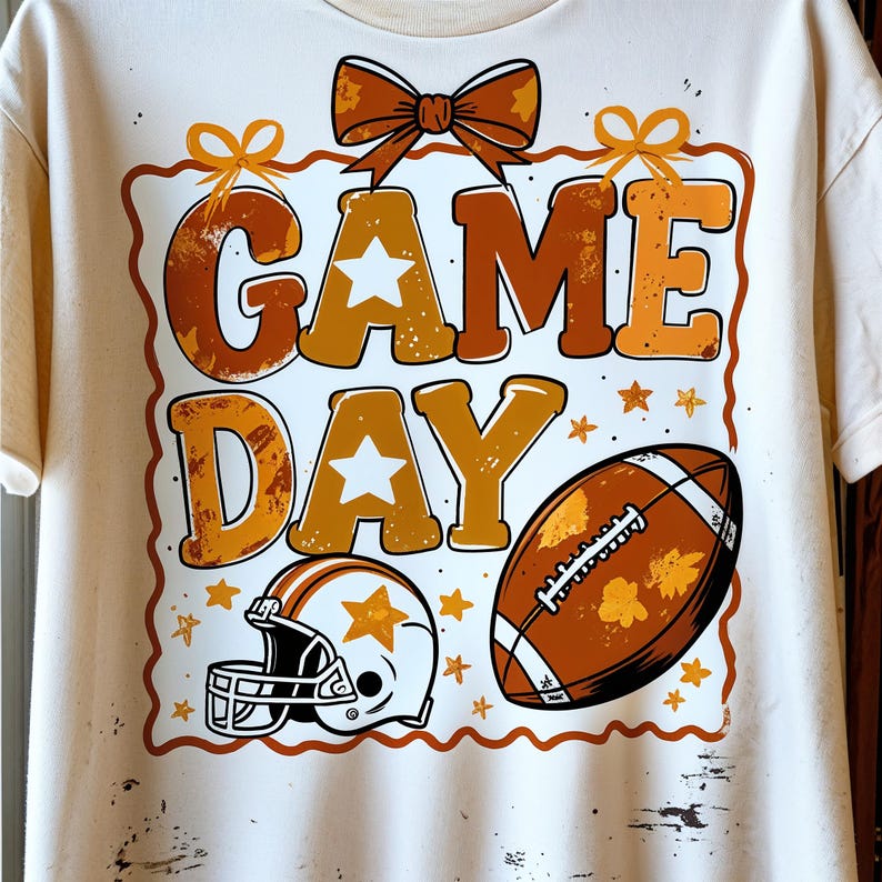 May include: Cream-colored t-shirt featuring a football theme. The design includes the words "GAME DAY" in orange and brown, a football, a helmet, and star accents. A bow and decorative border complete the design.