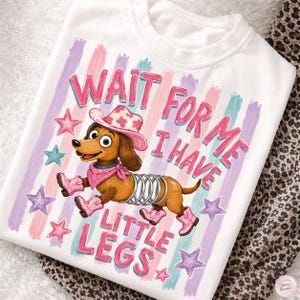 Puede incluir: Una camiseta blanca con un perro salchicha marrón con cuerpo de muelle, que lleva un sombrero de vaquero rosa, un pañuelo rosa y botas de vaquero rosas. El texto "WAIT FOR ME I HAVE LITTLE LEGS" está en rosa, rodeado de rayas y estrellas pastel.