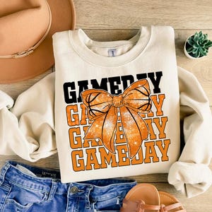 Może przedstawiać: Kremowa bluza z napisem "GAME DAY" w czarnych i pomarańczowych literach blokowych. Duża pomarańczowa kokarda, zaprojektowana tak, aby wyglądała jak koszyk&oacute;wka, jest wyśrodkowana z przodu. Bluza jest połączona z niebieskimi szortami dżinsowymi i sandałami.