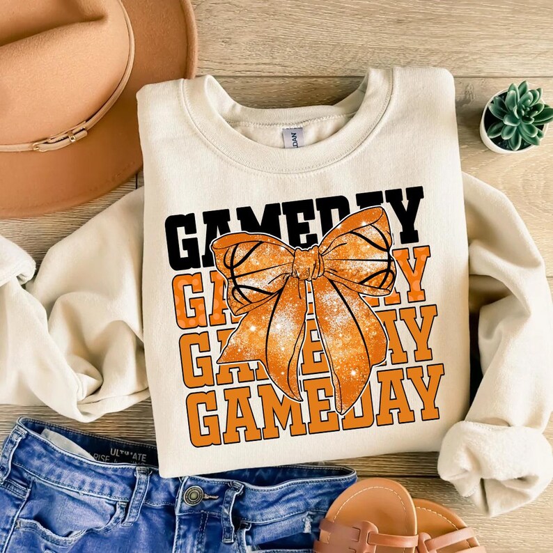 Może przedstawiać: Kremowa bluza z napisem "GAME DAY" w czarnych i pomarańczowych literach blokowych. Duża pomarańczowa kokarda, zaprojektowana tak, aby wyglądała jak koszyk&oacute;wka, jest wyśrodkowana z przodu. Bluza jest połączona z niebieskimi szortami dżinsowymi i sandałami.