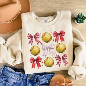 May include: A cream-colored sweatshirt with a pattern of red and white gingham bows and yellow softball illustrations. The softball illustrations have red stitching.