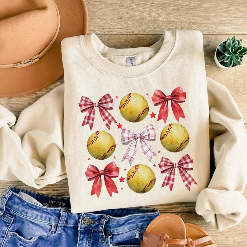May include: A cream-colored sweatshirt with a pattern of red and white gingham bows and yellow softball illustrations. The softball illustrations have red stitching.