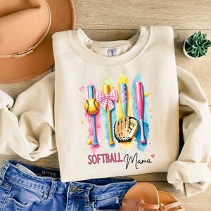 May include: A cream colored sweatshirt with a watercolor design of softball bats, a ball, a glove, and a pink bow. The text "Softball Mama" is printed below the design.