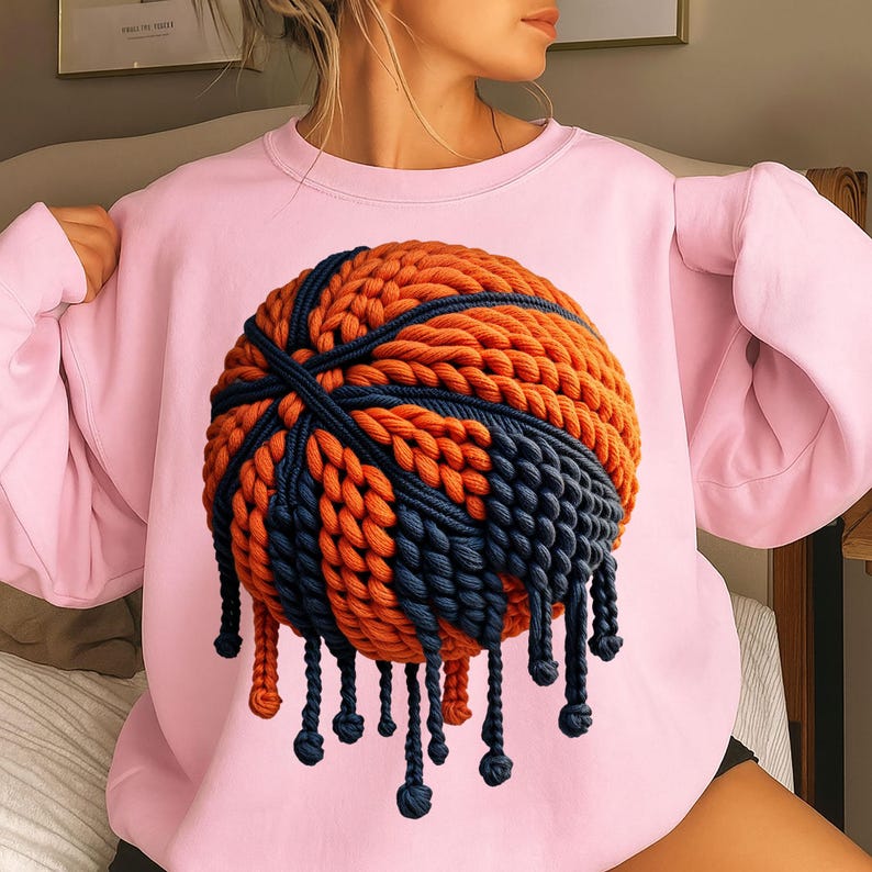 May include: A light pink sweatshirt featuring a basketball design. The basketball is rendered in orange and navy blue, with a textured, woven appearance and dangling fringe.
