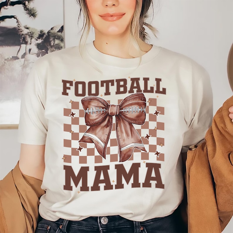 May include: Beige t-shirt with the words "FOOTBALL MAMA" in brown block letters. A brown football bow graphic is centered on a brown and white checkered background. The shirt is a casual, everyday wear item.