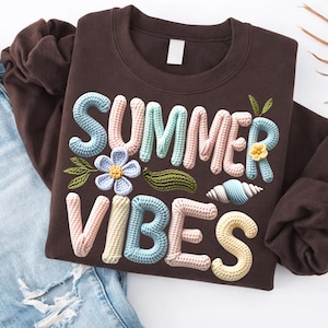 Może przedstawiać: Brązowa bluza z napisem "SUMMER VIBES" w tr&oacute;jwymiarowym wzorze szydełkowym. Projekt zawiera kwiat, liście i muszlę w pastelowych kolorach. Bluza jest złożona na niebieskich dżinsach.