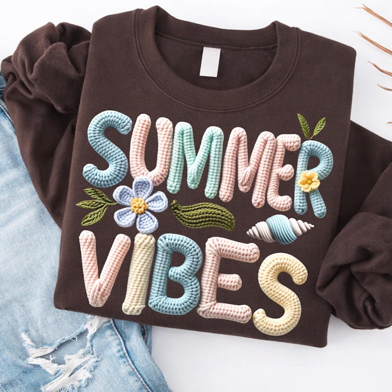 Może przedstawiać: Brązowa bluza z napisem "SUMMER VIBES" w tr&oacute;jwymiarowym wzorze szydełkowym. Projekt zawiera kwiat, liście i muszlę w pastelowych kolorach. Bluza jest złożona na niebieskich dżinsach.