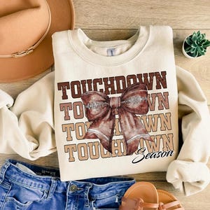 May include: Cream-colored sweatshirt with the words "TOUCHDOWN" in brown and tan. A football-shaped bow graphic is centered on the front. The word "Season" is written in script below the bow. The sweatshirt is paired with blue jeans and sandals.
