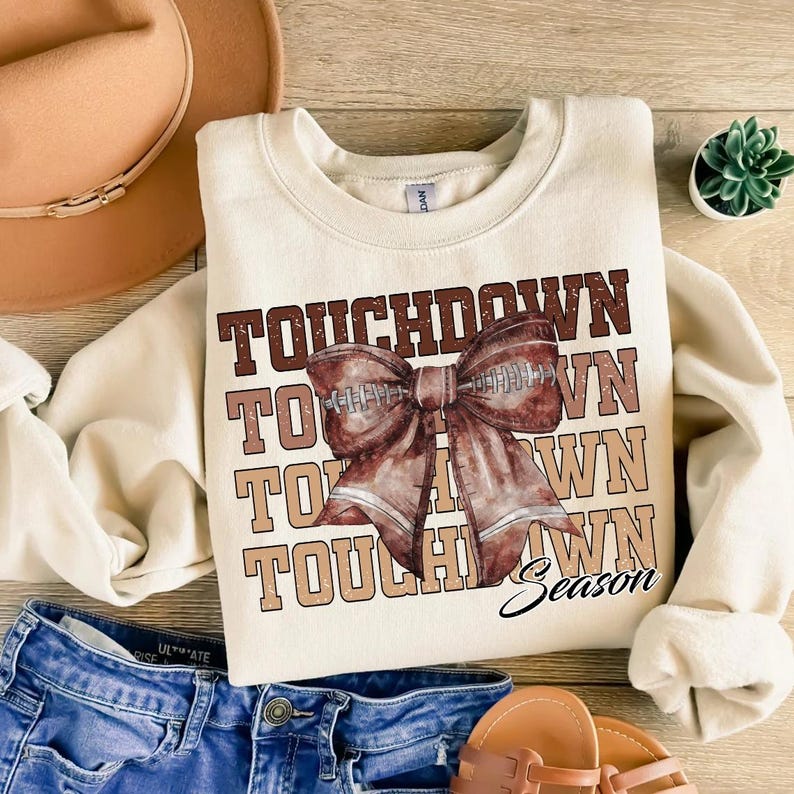 May include: Cream-colored sweatshirt with the words "TOUCHDOWN" in brown and tan. A football-shaped bow graphic is centered on the front. The word "Season" is written in script below the bow. The sweatshirt is paired with blue jeans and sandals.