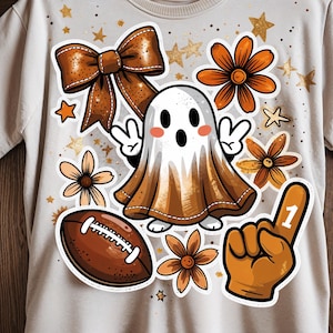 May include: A light-colored t-shirt featuring a collection of fall-themed stickers. The stickers include a brown bow, a friendly ghost, a football, orange flowers, a number one hand, and gold stars. The design has a festive, seasonal theme.