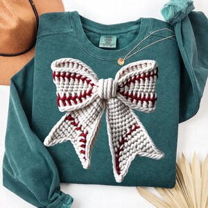 May include: Teal green sweatshirt featuring a large, textured bow design. The bow is primarily off-white with red stitching, creating a baseball-like pattern. A gold necklace and a brown hat are visible in the background. The sweatshirt has a crew neck and long sleeves.