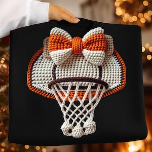 May include: Black tote bag featuring a crocheted basketball hoop design. The hoop is white and orange, with a brown rim and a white net. A large bow in matching colors sits above the hoop. The bag is held by a person.
