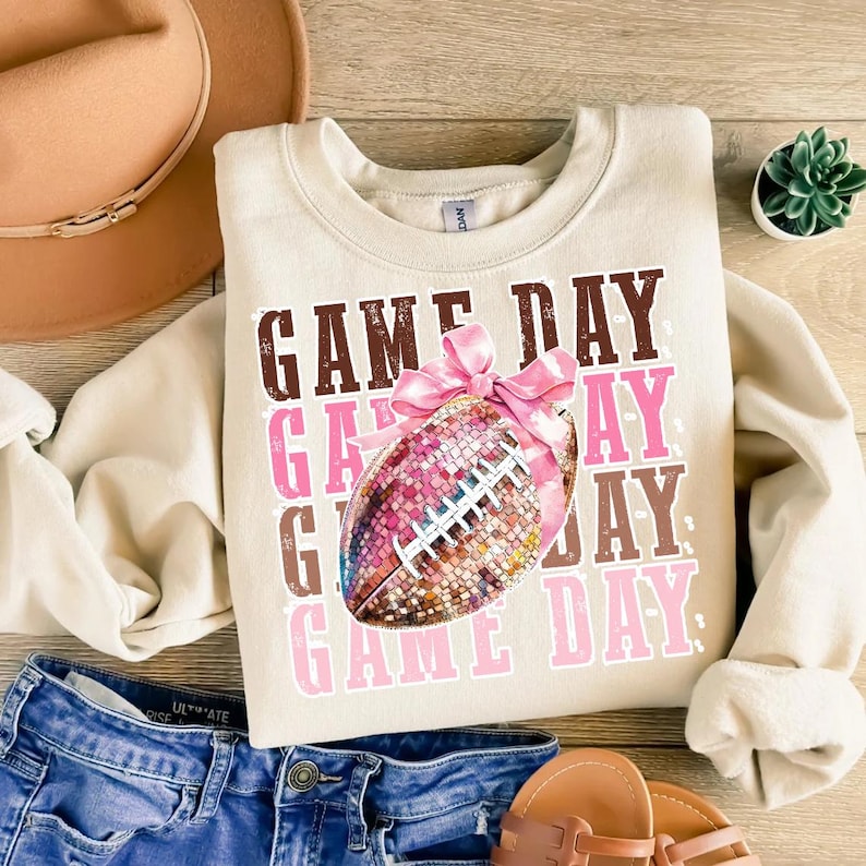May include: A beige sweatshirt with the words "GAME DAY" in brown and pink. A football with a pink bow is in the center. The sweatshirt is paired with blue denim shorts, a brown hat, and sandals.