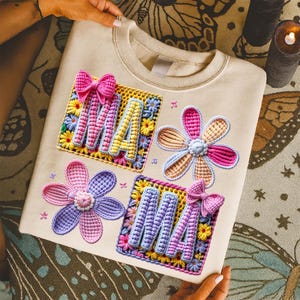May include: Cream-colored sweatshirt with colorful embroidered floral and "MA" designs. The "MA" letters are in a square with a bow, and the flowers are in shades of pink, purple, and yellow. The sweatshirt is held by hands.
