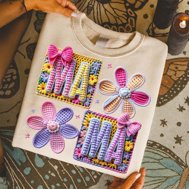 May include: Cream-colored sweatshirt with colorful embroidered floral and "MA" designs. The "MA" letters are in a square with a bow, and the flowers are in shades of pink, purple, and yellow. The sweatshirt is held by hands.