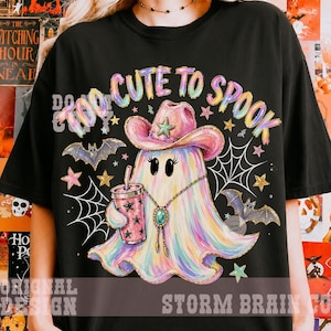May include: A black t-shirt featuring a colorful, iridescent ghost wearing a pink cowboy hat and a turquoise bolo tie. The ghost holds a pink star-patterned cup. The text reads TOO CUTE TO SPOOK, surrounded by bats, spiderwebs, and stars.