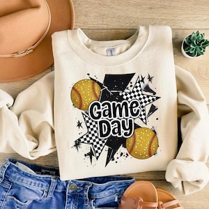 May include: A cream-colored sweatshirt with a black and white checkered star design, two yellow softball graphics, and the text "Game Day".