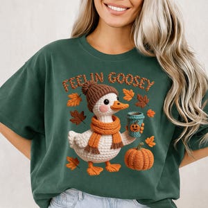 May include: Green t-shirt with a fall-themed graphic. The graphic features a crocheted duck wearing a brown hat and orange scarf, holding a coffee cup with a pumpkin face. The text "FEELIN GOOSEY" is above the duck, with fall leaves and a pumpkin.