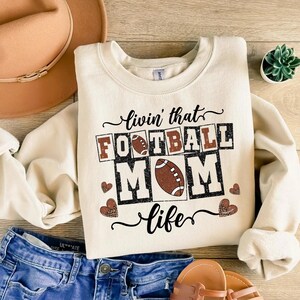 May include: Cream-colored sweatshirt with the text "livin' that FOOTBALL MOM life" in black and brown. The design includes footballs and heart shapes with a leopard print pattern. The sweatshirt is paired with blue jeans and a brown hat.