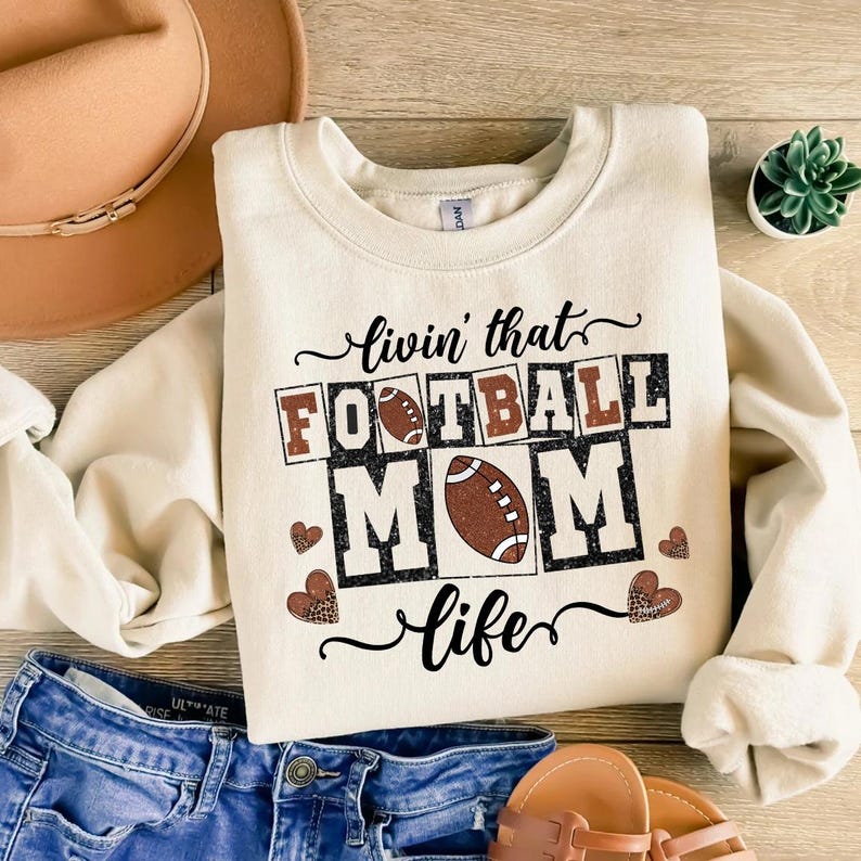 May include: Cream-colored sweatshirt with the text "livin' that FOOTBALL MOM life" in black and brown. The design includes footballs and heart shapes with a leopard print pattern. The sweatshirt is paired with blue jeans and a brown hat.
