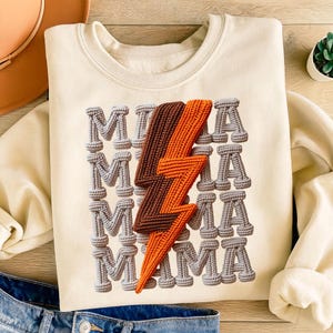 May include: Cream-colored sweatshirt with the word "MAMA" repeated vertically in gray, rope-like letters. A brown and orange lightning bolt design is centered on the front. The sweatshirt is folded, with a portion of blue jeans visible.