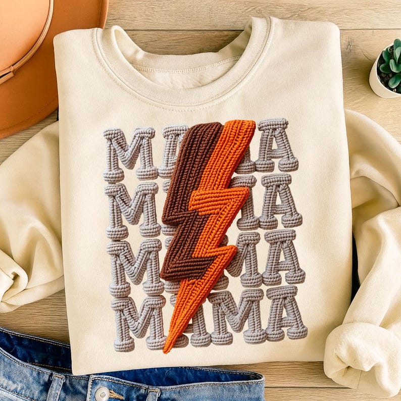 May include: Cream-colored sweatshirt with the word "MAMA" repeated vertically in gray, rope-like letters. A brown and orange lightning bolt design is centered on the front. The sweatshirt is folded, with a portion of blue jeans visible.