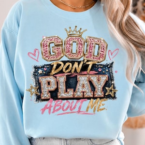 May include: A light blue crewneck sweatshirt featuring a graphic print with the text "GOD DON'T PLAY ABOUT ME". The design includes pink glittery letters, a gold crown, gold stars, and pink hearts, all set against a dark blue denim-textured background.