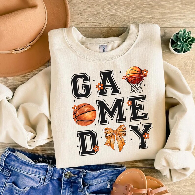 Może przedstawiać: Kremowa bluza z napisem "GAME DAY" w czarnych literach blokowych, ozdobiona koszyk&oacute;wkami, siatką i kokardą. Pomarańczowe akcenty kwiatowe są rozrzucone wok&oacute;ł tekstu. Bluza jest połączona z niebieskimi szortami dżinsowymi i sandałami.