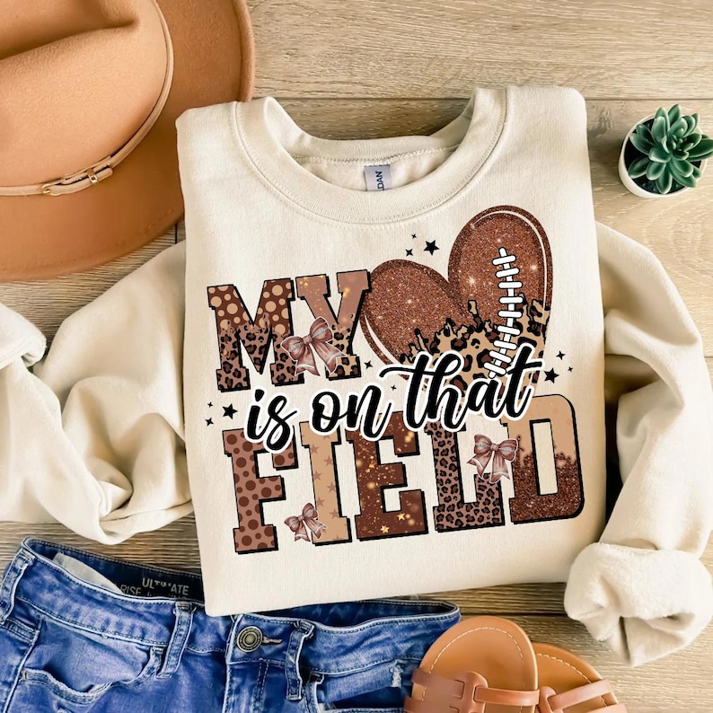 May include: Cream-colored sweatshirt with the text "MY is on that FIELD" in brown and leopard print. A football-shaped heart is in the center. The sweatshirt is paired with blue denim shorts and sandals.