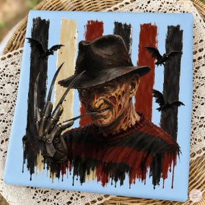 May include: A square canvas print featuring a portrait of a character with a scarred face, wearing a brown fedora and a red and black striped sweater. The character has a metal clawed glove on one hand, with black birds flying against a striped background.