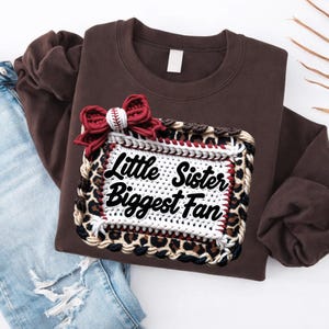 May include: A brown sweatshirt with a decorative patch that reads "Little Sister Biggest Fan." The patch has a leopard print border and a baseball-themed bow. The sweatshirt is paired with blue jeans.