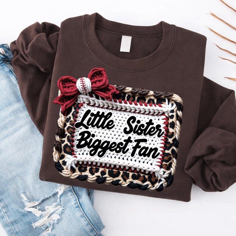May include: A brown sweatshirt with a decorative patch that reads "Little Sister Biggest Fan." The patch has a leopard print border and a baseball-themed bow. The sweatshirt is paired with blue jeans.