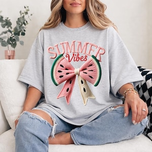 Może przedstawiać: Jasnoszara koszulka oversize z motywem kokardy z arbuza. Kokarda jest r&oacute;żowa, zielona i beżowa, z napisem "SUMMER VIBES" w kolorze r&oacute;żowym i zielonym powyżej. Koszulka jest wykonana z miękkiego materiału.