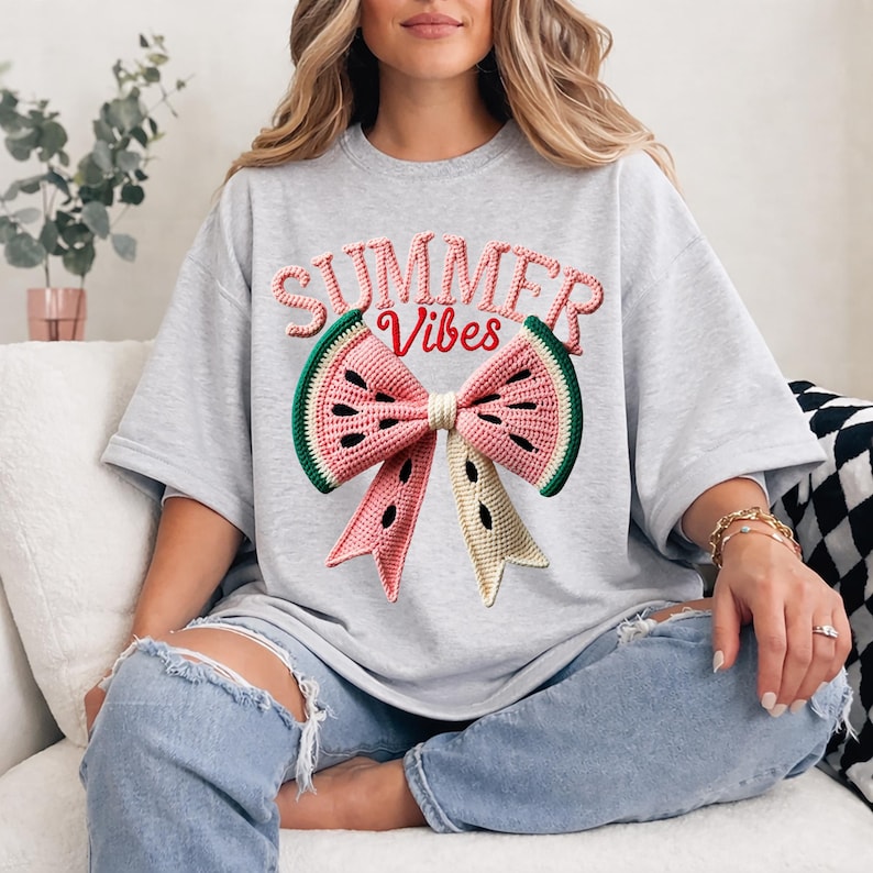 Może przedstawiać: Jasnoszara koszulka oversize z motywem kokardy z arbuza. Kokarda jest r&oacute;żowa, zielona i beżowa, z napisem "SUMMER VIBES" w kolorze r&oacute;żowym i zielonym powyżej. Koszulka jest wykonana z miękkiego materiału.