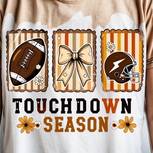 May include: White t-shirt with a football-themed graphic. The design includes a football, a bow, and a football helmet, all within decorative frames. The text "TOUCHDOWN SEASON" is displayed below, with floral accents.