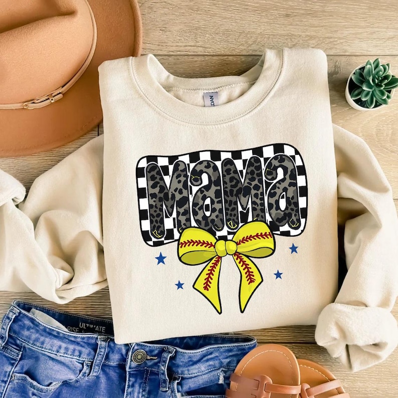 May include: A cream-colored sweatshirt with a black and white checkered design featuring the word "Mama" in a leopard print. A yellow softball bow with red stitching is centered below the word "Mama".
