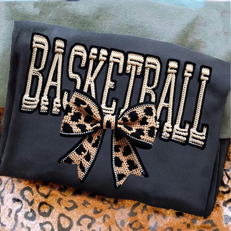 May include: Dark gray sweatshirt with the word "BASKETBALL" in cream-colored, embroidered letters. A leopard print bow is centered below the text. The sweatshirt is folded on a patterned surface.
