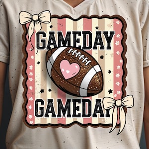 May include: A beige t-shirt with a football-themed graphic. The graphic features a brown football with a pink heart, the text "GAMEDAY" in black, and decorative bows and stars. The design has a vintage, slightly distressed look.