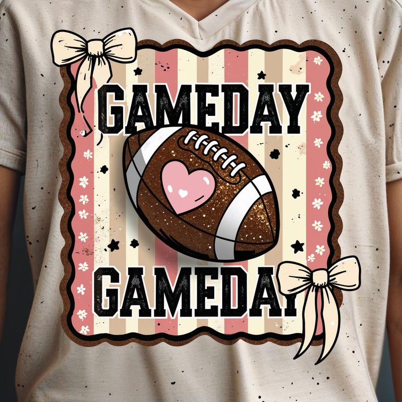May include: A beige t-shirt with a football-themed graphic. The graphic features a brown football with a pink heart, the text "GAMEDAY" in black, and decorative bows and stars. The design has a vintage, slightly distressed look.