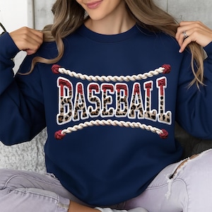 May include: Navy blue sweatshirt with the word "BASEBALL" in a bold, stylized font. The letters have a leopard print pattern and are outlined in white. The word is framed by a white rope design with red accents.
