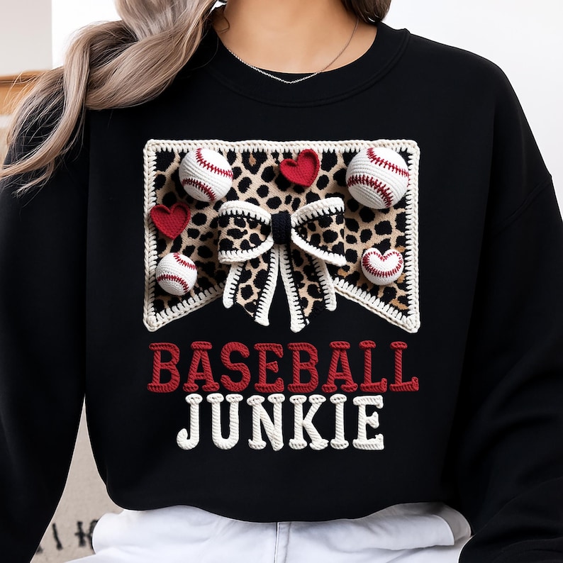 May include: Black sweatshirt with a baseball-themed design. The design includes baseballs, red hearts, and a leopard print bow. The words "BASEBALL JUNKIE" are embroidered in red and white.