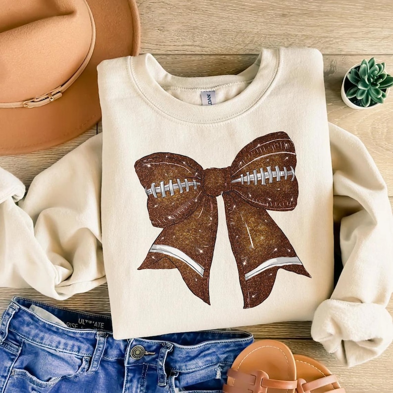 May include: Cream-colored sweatshirt featuring a large, glittery brown bow designed to resemble a football. The bow has white stripes and stitching details. The sweatshirt is part of a casual apparel collection.