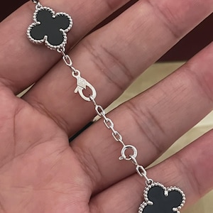 May include: A silver-toned bracelet featuring two black clover-shaped charms with beaded edges. The charms are connected by a silver chain link segment that includes a lobster claw clasp and a circular spring ring clasp for secure fastening.