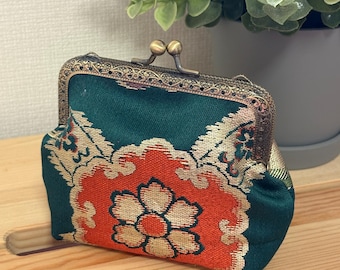 Puede incluir: Un monedero pequeño con base de tela verde oscuro y un llamativo patrón floral en naranja y dorado. El monedero se asegura con un cierre de boquilla de metal bronce con estilo antiguo, que presenta intrincados detalles de filigrana.