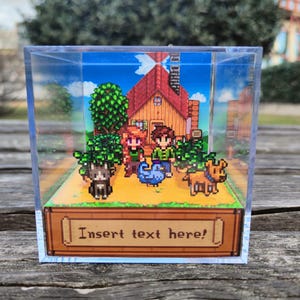 Könnte beinhalten: Ein klarer, würfelförmiger Display mit einer pixeligen Szene mit einem Haus, Figuren und Tieren. Die Basis hat ein Banner mit der Aufschrift "Insert text here!". Die Szene spielt vor blauem Himmel.