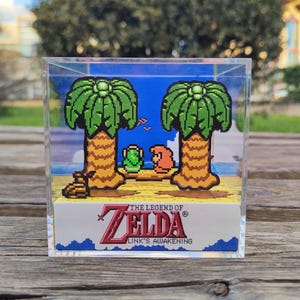 Puede incluir: Un cubo transparente con un diorama 3D de una escena de The Legend of Zelda: Link's Awakening. El diorama presenta a Link y una criatura verde de pie en una playa con dos palmeras. El cubo tiene una base blanca con el título y el logotipo del juego.