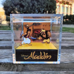 Puede incluir: Una caja de acrílico transparente con un diorama 3D de Aladdin y Jafar del videojuego Disney's Aladdin. El diorama está sobre un fondo amarillo con el texto "Disney's Aladdin" en la parte inferior.
