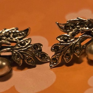 May include: A pair of silver-toned leaf-shaped earrings with marcasite accents and a faux pearl at the base. The earrings are set against an orange background.