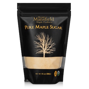 May include: A black resealable pouch with a label that reads "Mount Mansfield Maple Products Pure Maple Sugar". The label also features a stylized maple tree and the text "Net Wt 14 oz (396g)". The pouch contains a light brown maple sugar.